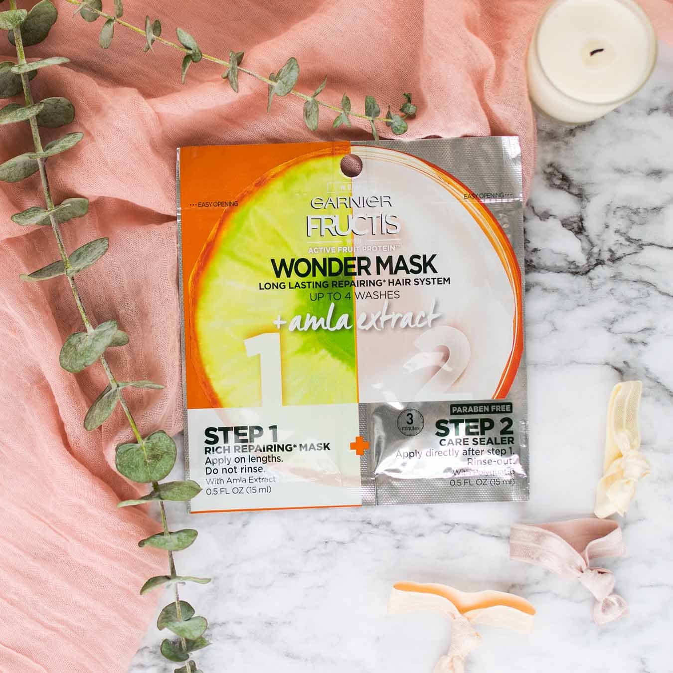 Garnier Fructis Wonder Mask with Amla Extract on white marble and pink linen next to a white candle, colored knotted elastics, and two eucalyptus branches with tiered leaves.